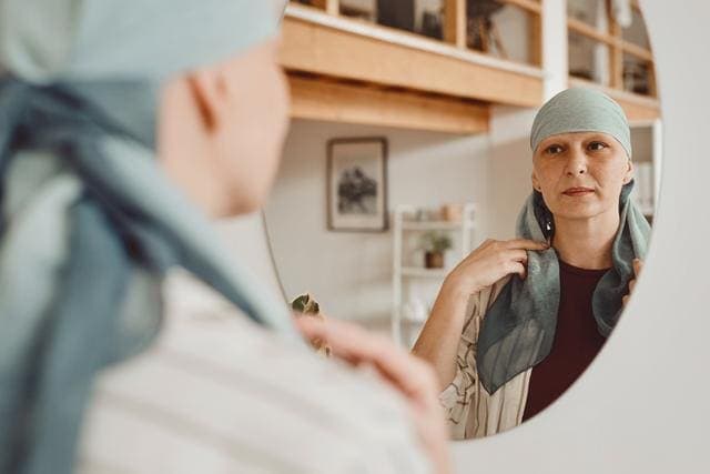 Woman putting on a head scarf while looking in the mirror standing in home interior, alopecia and cancer awareness.